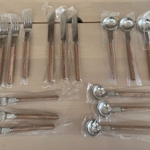 May include: A collection of stainless steel silverware with wooden handles, including forks, knives, and spoons. The utensils are individually wrapped in clear plastic, ready for use. The wooden handles have a light brown color.
