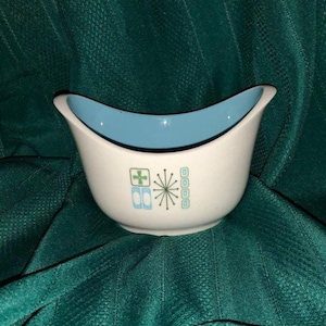 May include: A white ceramic bowl with a light blue interior. The bowl has a unique, curved shape and is decorated with a retro-style design in blue and green. The design includes a cross, a starburst, and three circles.