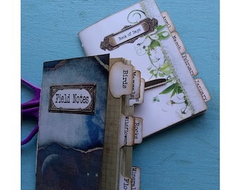May include: Two small, handmade journals with decorative covers. The journal on the left has a blue and white cover with the words "Field Notes" on it. The journal on the right has a white cover with the words "Book of Days" on it. Both journals have tabs with different categories written on them, such as "Birds", "Rocks", and "Flowers".