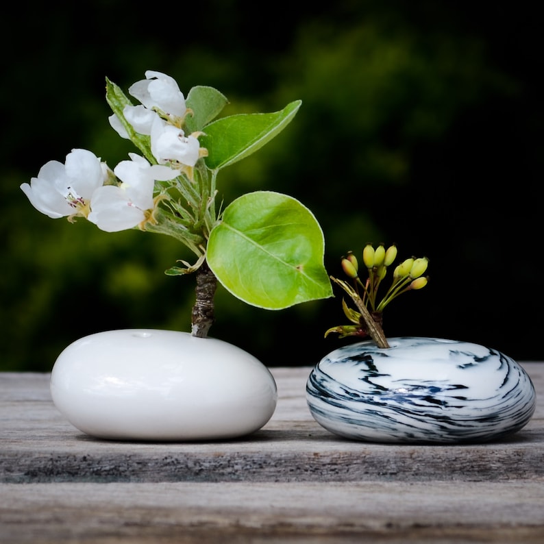 May include: Two white ceramic vases with a simple, modern design. One vase holds a small arrangement of white blossoms and green leaves, while the other vase holds a small arrangement of green leaves.