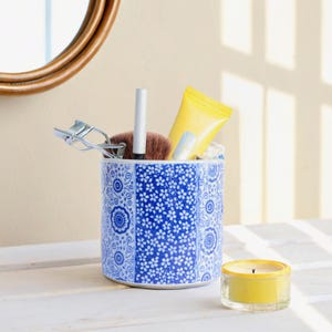 Può includere: Un contenitore in ceramica blu e bianca con un motivo floreale contiene pennelli per il trucco, un tubo di crema solare e un piegaciglia. Una candela gialla si trova su una superficie bianca.