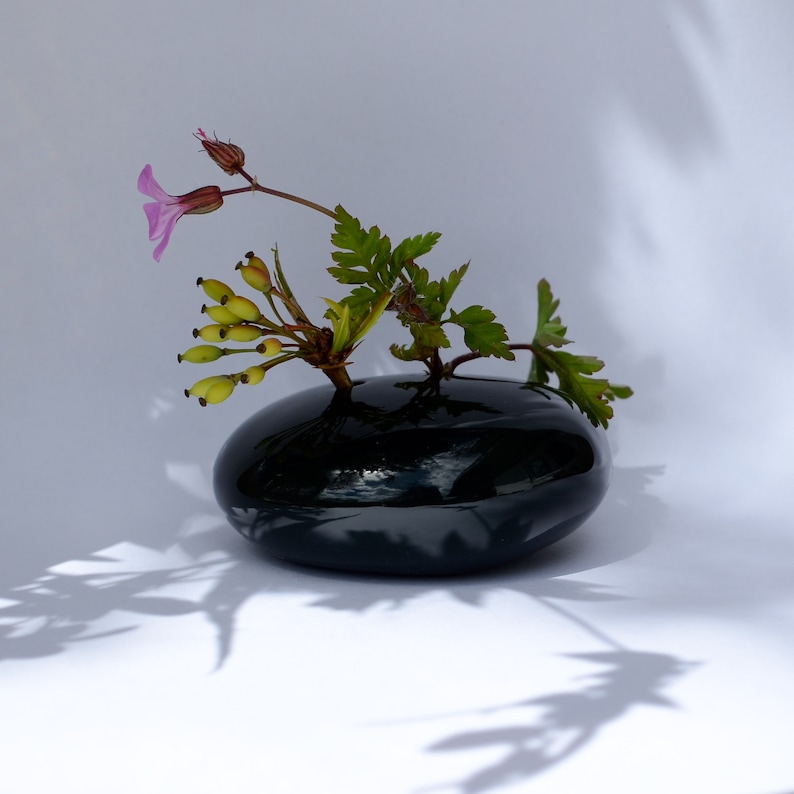 May include: A small arrangement of pink flowers and green leaves in a black, round vase. The vase is sitting on a white surface.