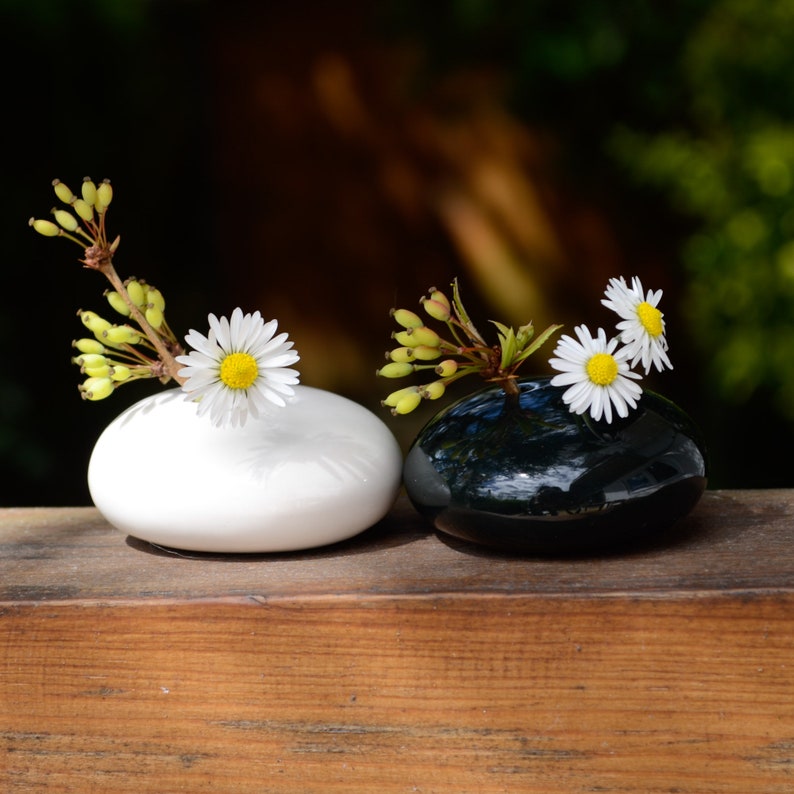 May include: Two small, round vases, one white and one black, each holding a single white daisy. The vases are sitting on a wooden surface.