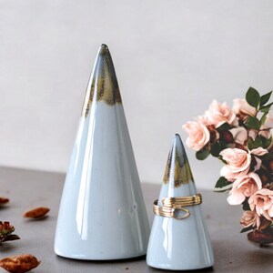 May include: Two ceramic ring holders shaped like mountains. The larger holder is light blue with a brown glaze at the top. The smaller holder is light blue with a brown glaze at the top and a gold ring on it.