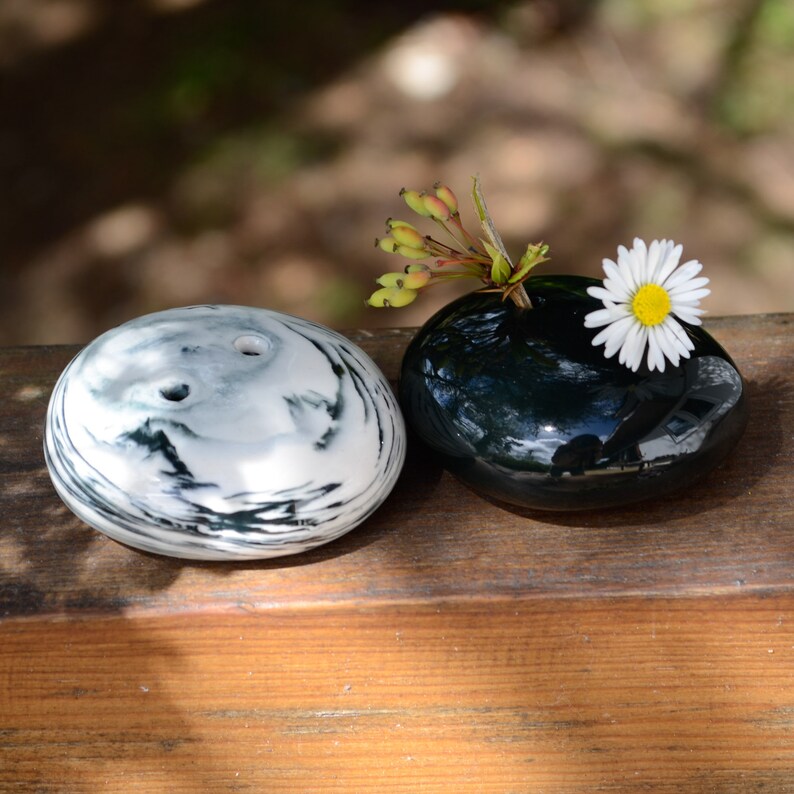 May include: Two small ceramic vases, one black and one white with black swirls, on a wooden surface. The black vase has a single white daisy with green stems.
