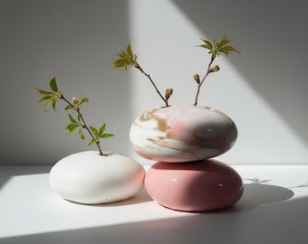 May include: Three ceramic vases in a stack, each with a rounded, pebble-like shape. The bottom vase is a solid pink, the middle one is white with pink and brown marbling, and the top one is white. Each vase holds a small sprig of greenery.