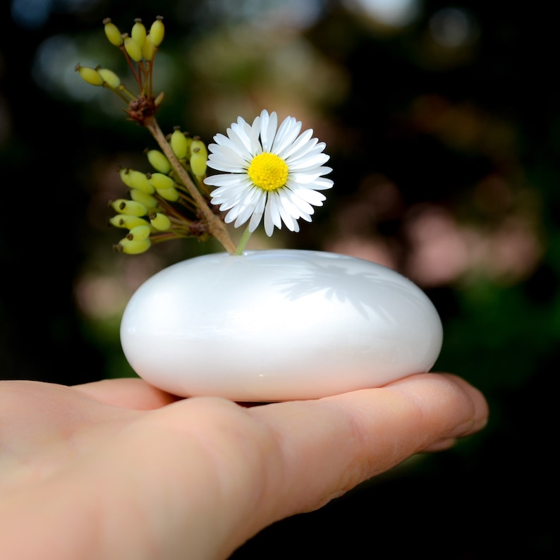 May include: A single white daisy with a yellow center is placed in a small white ceramic vase. The vase is shaped like a smooth, rounded stone. The vase is held in a person's hand.