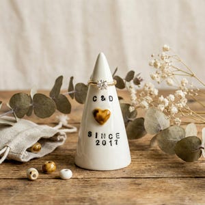 Puede incluir: Un soporte para anillos de cerámica blanca en forma cónica, con un anillo de oro en la parte superior. El soporte tiene las iniciales "C & D", un corazón dorado y el texto "SINCE 2017". El fondo incluye flores secas y vegetación.