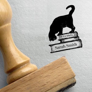 K&ouml;nnte beinhalten: Eine schwarze Katze mit einem eingerollten Schwanz steht auf drei &uuml;bereinander gestapelten B&uuml;chern. Das oberste Buch tr&auml;gt den Text "Ex libris" aufgedruckt. Das unterste Buch tr&auml;gt den Text "Sarah Smith" aufgedruckt. Das Bild ist ein Stempel.