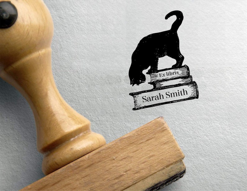 K&ouml;nnte beinhalten: Eine schwarze Katze mit einem eingerollten Schwanz steht auf drei &uuml;bereinander gestapelten B&uuml;chern. Das oberste Buch tr&auml;gt den Text "Ex libris" aufgedruckt. Das unterste Buch tr&auml;gt den Text "Sarah Smith" aufgedruckt. Das Bild ist ein Stempel.
