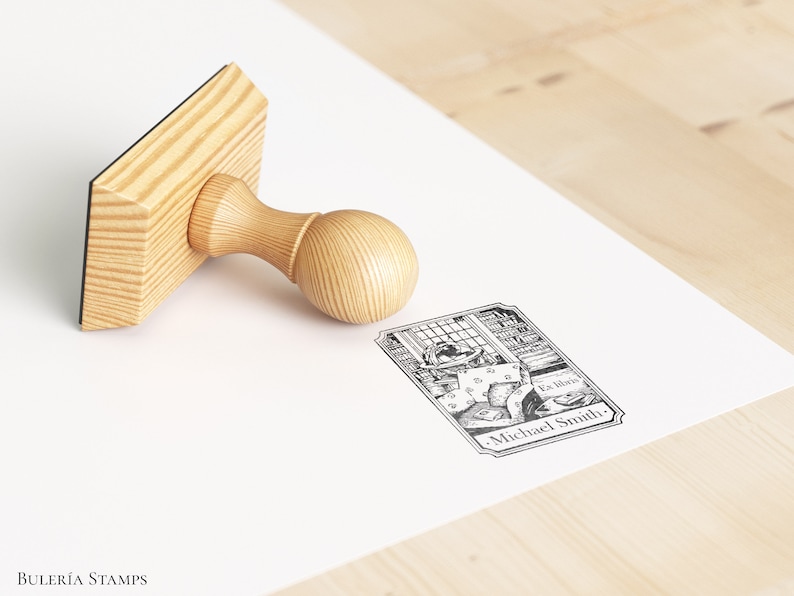 May include: A wooden stamp with a handle and a rectangular base. The stamp features a black and white illustration of a person reading in a library with the text "La libra" and "Michael Smith".