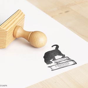 K&ouml;nnte beinhalten: Ein Stempel aus Holz mit einem schwarzen Stempelkissen, auf dem eine schwarze Katzensilhouette auf einem Stapel B&uuml;cher sitzt. Der Text "Los libros" und "Cristine Pastrana" ist auf den B&uuml;chern gedruckt. Der Stempel befindet sich auf einer wei&szlig;en Oberfl&auml;che.