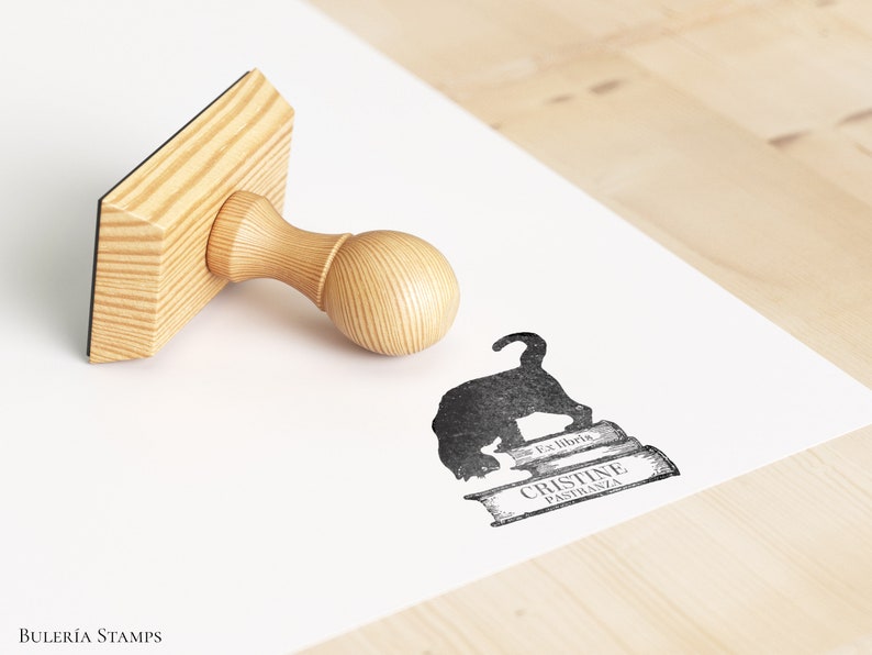 K&ouml;nnte beinhalten: Ein Stempel aus Holz mit einem schwarzen Stempelkissen, auf dem eine schwarze Katzensilhouette auf einem Stapel B&uuml;cher sitzt. Der Text "Los libros" und "Cristine Pastrana" ist auf den B&uuml;chern gedruckt. Der Stempel befindet sich auf einer wei&szlig;en Oberfl&auml;che.