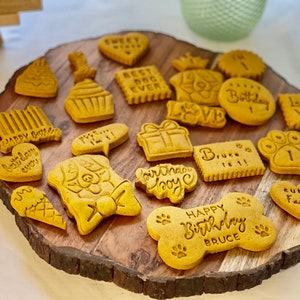 May include: A wooden platter with a variety of dog-themed birthday cookies. The cookies are decorated with different messages, including "Happy Birthday", "Best Dog Ever", "Love", and "Bruce is 1".