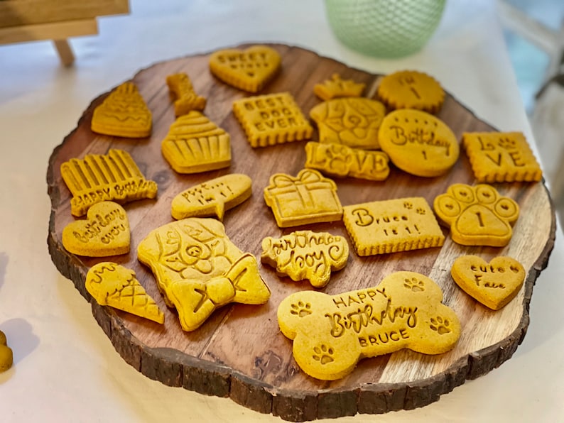 May include: A wooden platter with a variety of dog-themed birthday cookies. The cookies are decorated with different messages, including "Happy Birthday", "Best Dog Ever", "Love", and "Bruce is 1".