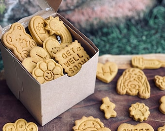 Op de afbeelding: Een kartonnen doos gevuld met diverse hondenkoekjes. De koekjes hebben verschillende vormen, waaronder een beer, een pootafdruk, een bloem en een hart. Eén koekje heeft de tekst "GET WELL SOON". De koekjes zijn lichtbruin en liggen op een houten oppervlak.