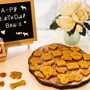 May include: A birthday celebration for a dog named Bruce with a chalkboard sign that says "Happy Birthday Bruce", a box of dog treats, and a variety of dog biscuits on a wooden platter. The biscuits are decorated with various dog-themed designs and messages, including "Happy Birthday Bruce", "Best Dog Ever", and "Sweet Treats".