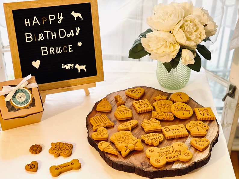 May include: A birthday celebration for a dog named Bruce with a chalkboard sign that says "Happy Birthday Bruce", a box of dog treats, and a variety of dog biscuits on a wooden platter. The biscuits are decorated with various dog-themed designs and messages, including "Happy Birthday Bruce", "Best Dog Ever", and "Sweet Treats".