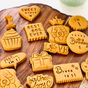 May include: A variety of dog birthday cookies on a wooden surface. The cookies are decorated with different designs, including a cupcake, a present, a dog's face, and the words "Best Dog Ever", "Sweet Bruce", "I'm 1", "Birthday Boy", and "Bruce is 1!"