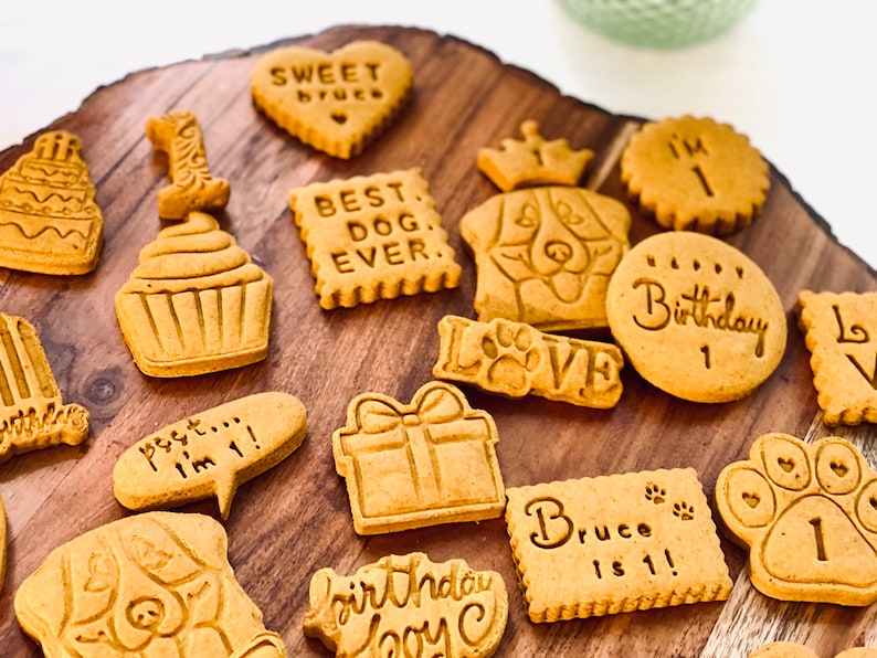May include: A variety of dog birthday cookies on a wooden surface. The cookies are decorated with different designs, including a cupcake, a present, a dog's face, and the words "Best Dog Ever", "Sweet Bruce", "I'm 1", "Birthday Boy", and "Bruce is 1!"