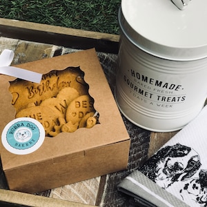 May include: A brown box with a clear window showing dog biscuits with the words "Happy Birthday" and "We Love Dogs" on them. The box has a sticker that says "Bubba Dog Bakery". A white ceramic jar with a silver paw print lid says "Homemade. Baked Fresh. Gourmet Treats. 7 Days A Week". A white and gray striped kitchen towel with a black and white illustration of two dogs is on a wooden tray.