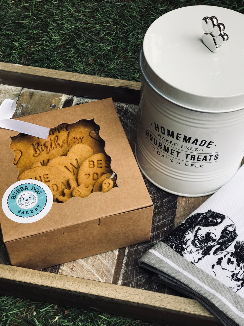 May include: A brown box with a clear window showing dog biscuits with the words "Happy Birthday" and "We Love Dogs" on them. The box has a sticker that says "Bubba Dog Bakery". A white ceramic jar with a silver paw print lid says "Homemade. Baked Fresh. Gourmet Treats. 7 Days A Week". A white and gray striped kitchen towel with a black and white illustration of two dogs is on a wooden tray.