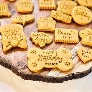 May include: A collection of dog-shaped biscuits decorated with various messages, including "Happy Birthday Bruce", "Birthday Boy", "Cute Face", and "Bruce is 1!".