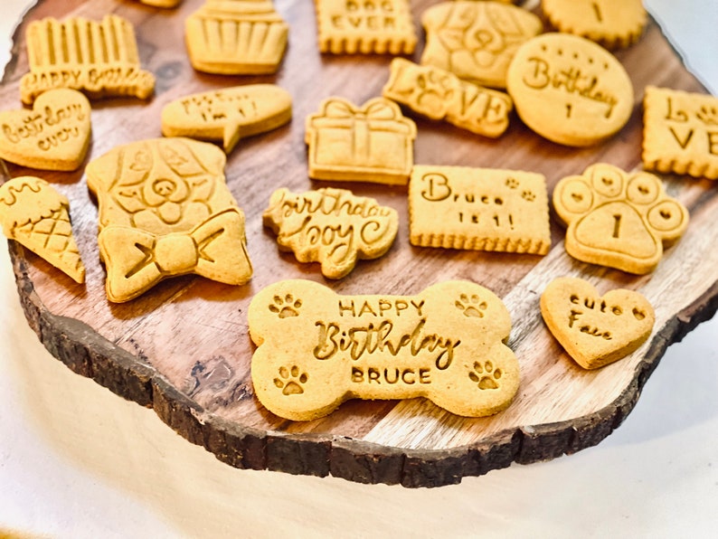 May include: A collection of dog-shaped biscuits decorated with various messages, including "Happy Birthday Bruce", "Birthday Boy", "Cute Face", and "Bruce is 1!".
