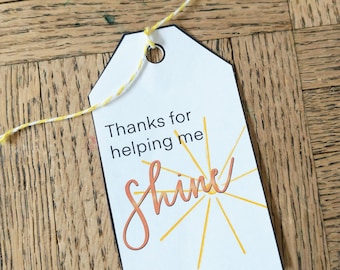 Op de afbeelding: Witte cadeaukaart met zwarte rand en zwarte tekst die "Bedankt dat je me helpt te stralen" zegt. De tekst is in een handschrift. Achter de tekst is een gele zon met stralen getekend.