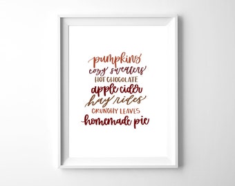 May include: A framed print with a white border features fall-themed words in a gradient of orange and red. The words include "pumpkins," "cozy sweaters," "hot chocolate," "apple cider," "hay rides," "crunchy leaves," and "homemade pie."