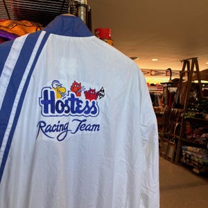 May include: Vintage white and blue racing jacket with "Hostess Racing Team" embroidered on the back. The jacket has blue stripes on the sleeves and a blue collar. The Hostess logo features cartoon characters.