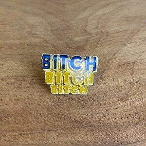 May include: A gold-coloured enamel pin featuring the word "BITCH" in blue and yellow. The word is stacked three times, with the top word in a bold, outlined font.