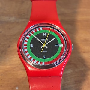 Peut inclure: Une montre Swatch rouge avec un cadran noir présentant un design de boussole avec des accents verts et blancs. La montre a un bracelet rouge et une lunette argentée. Le cadran affiche le logo Swatch Quartz et la date.