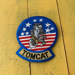 Könnte beinhalten: Gestickter Aufnäher mit einer Cartoon-Katze im militärischen Design. Der Aufnäher hat einen blauen Rand mit weißen Sternen, rot-weißen Streifen und dem Wort "TOMCAT" in goldfarbenen Buchstaben.