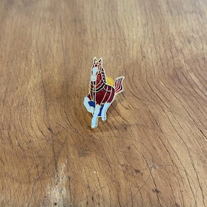 May include: A detailed enamel pin of a running horse. The horse has a red body with gold accents, a white face, and blue legs. The pin is set against a wooden background, highlighting the vibrant colours and intricate design of the horse.