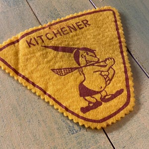 Peut inclure: Fanion en feutre jaune avec une bordure marron et le mot "KITCHENER" en haut. Un personnage de dessin animé avec un chapeau et une raquette est au centre. Le fanion a un bord festonné.