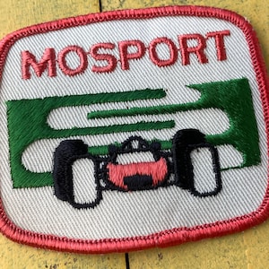 以下が含まれることがあります： 赤色の文字で「MOSPORT」と書かれた刺繍パッチ。緑と黒のレーシングカーのデザインが特徴です。パッチは白い背景と赤い縁取りが施されています。