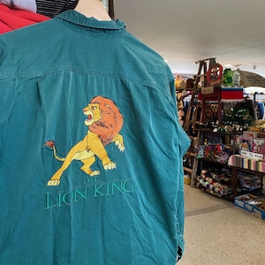 Könnte beinhalten: Türkisfarbenes Button-up-Shirt mit einem gestickten Bild von Simba aus Der König der Löwen. Das Bild zeigt Simba in einer Laufpose mit goldener Mähne und Körper. Der Text "THE LION KING" ist darunter in Grün gestickt.