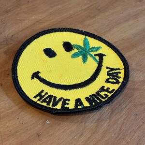 Peut inclure: Écusson jaune avec un visage souriant, des yeux noirs et une bouche incurvée. Une feuille de cannabis verte est sur le côté droit du visage. Les mots "HAVE A NICE DAY" sont brodés en noir en bas de l'écusson.