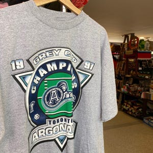 May include: A grey t-shirt featuring a blue and green graphic of the Toronto Argonauts 1997 Grey Cup Champions. The graphic depicts a football player wearing a helmet and the team's logo.