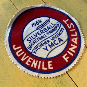 以下が含まれることがあります： 白、赤、青のデザインが施された、ヴィンテージの円形パッチ。「JUVENILE FINALIST」の文字が外縁に書かれています。中央には「1964 SILVERBALL BASKETBALL TOURNAMENT KITCHENER WATERLOO YMCA」の文字が書かれています。