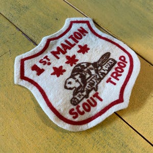 May include: A vintage, shield-shaped patch with a red border and text. The patch reads "1st Malton Scout Troop" in red, with a brown turtle illustration. The background is off-white.
