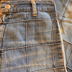 May include: Close-up of patchwork denim jeans. The jeans are a light blue wash with a grid-like pattern created by tan stitching. The jeans have a button closure and belt loops.