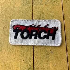 May include: A rectangular white patch featuring the word "TORCH" in black, bold lettering, with three red flames above. The patch is edged with white stitching. The background is a yellow wooden surface.