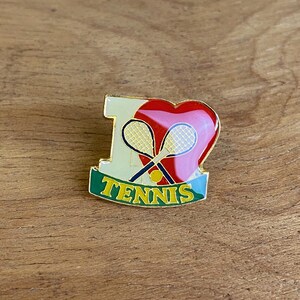 May include: A gold-coloured pin with the words "I heart Tennis". The pin features a red heart, two crossed tennis rackets, and a yellow tennis ball. The word "TENNIS" is written in gold letters on a green banner.