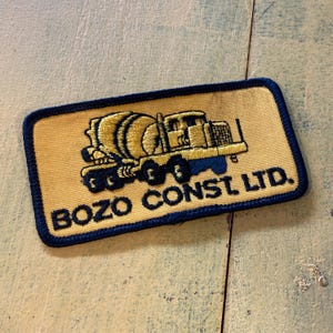 May include: A rectangular patch with a yellow background and a blue border. The patch features a yellow and blue illustration of a cement mixer truck. The text "BOZO CONST. LTD." is printed in blue.
