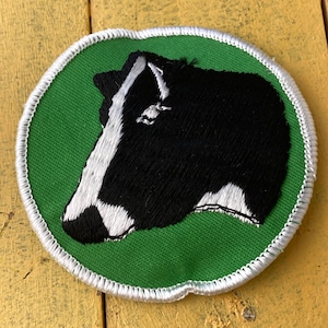 Op de afbeelding: Geborduurde patch met een zwart-witte koeienkop op een groene achtergrond. De patch heeft een wit gestikte rand. De koeienkop is in profiel afgebeeld, met gedetailleerde borduursels. De patch is rond.