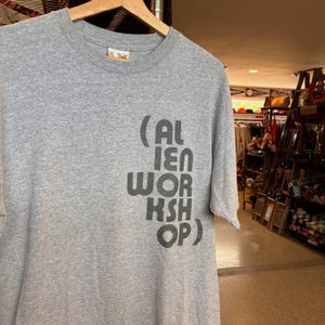 May include: A heather gray t-shirt with the text "(AL IEN WORK KSH OP)" printed in dark gray. The shirt is hanging on a wooden hanger. The background shows a store interior with various items.