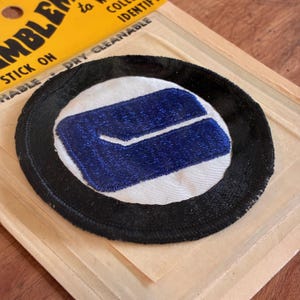 Peut inclure: Un écusson rond avec une bordure noire, un centre blanc et un motif brodé bleu. L'écusson est sur un support en carton avec du texte jaune.
