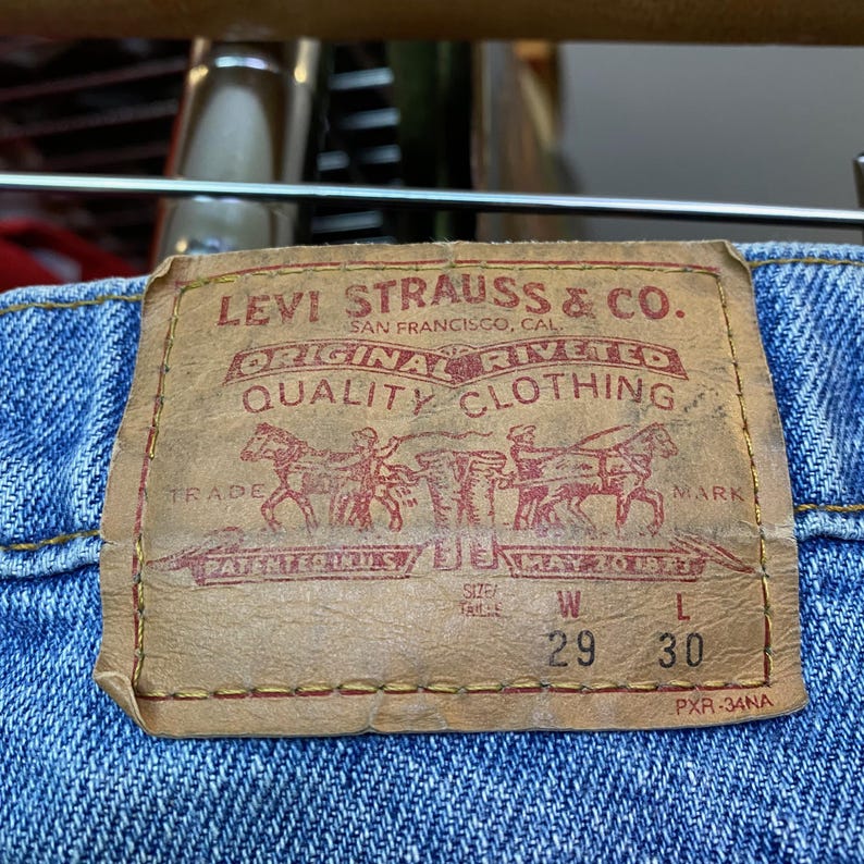 May include: Close-up of a Levi Strauss & Co. label on blue denim jeans. The label is tan with red lettering and a graphic of people and horses. The jeans are size W29 L30.
